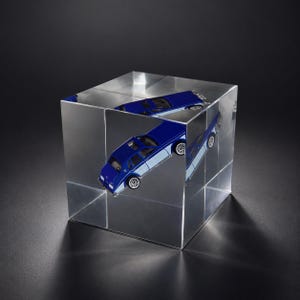 Puede incluir: Un cubo de acrílico transparente que contiene un coche de juguete en miniatura de color azul y azul claro. El coche está colocado en diagonal dentro del bloque, creando múltiples reflejos de la carrocería, las ruedas y las ventanas interiores.