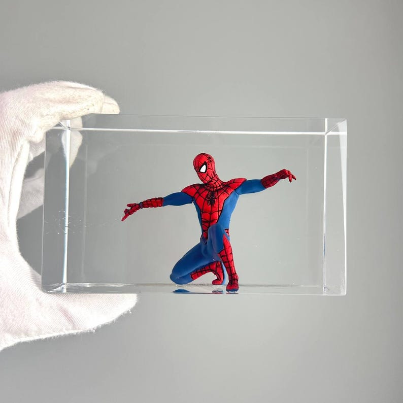 Peut inclure: Figurine de Spider-Man dans un pr&eacute;sentoir en acrylique transparent. La figurine est agenouill&eacute;e, les bras tendus. Le costume est rouge et bleu avec un motif de toile d'araign&eacute;e noire sur le masque et la poitrine.