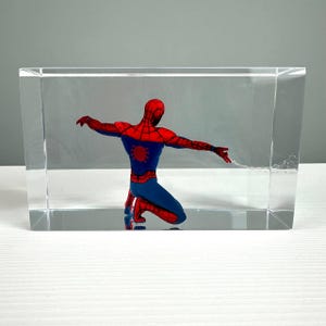 Peut inclure: Figurine de Spiderman agenouill&eacute;, enferm&eacute;e dans un bloc de r&eacute;sine transparent. Le super-h&eacute;ros porte son costume classique rouge et bleu, les bras tendus, avec une toile d'araign&eacute;e qui s'&eacute;tend de sa main. Le bloc est rectangulaire et repose sur une surface blanche.