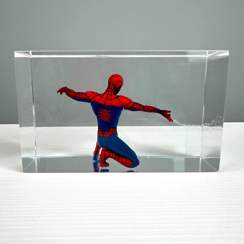 Peut inclure: Figurine de Spiderman agenouill&eacute;, enferm&eacute;e dans un bloc de r&eacute;sine transparent. Le super-h&eacute;ros porte son costume classique rouge et bleu, les bras tendus, avec une toile d'araign&eacute;e qui s'&eacute;tend de sa main. Le bloc est rectangulaire et repose sur une surface blanche.