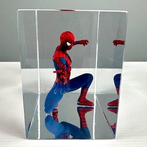 Peut inclure: Une figurine de Spider-Man rouge et bleu est enferm&eacute;e dans un bloc clair et rectangulaire. La figurine est en position accroupie, avec son reflet visible. Le bloc est sur une surface blanche.