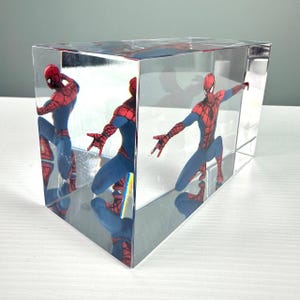 Peut inclure: Un bloc rectangulaire transparent enferme une figurine de Spider-Man dans une pose dynamique. Le super-h&eacute;ros est v&ecirc;tu d'un costume rouge et bleu, les bras tendus. Les c&ocirc;t&eacute;s miroit&eacute;s du bloc refl&egrave;tent la figurine, cr&eacute;ant plusieurs vues.