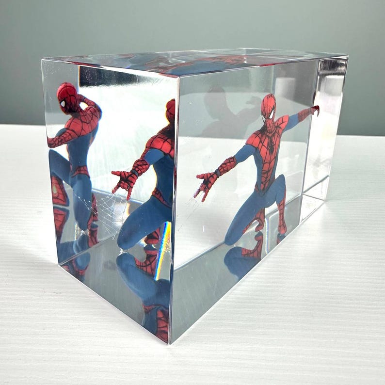 Peut inclure: Un bloc rectangulaire transparent enferme une figurine de Spider-Man dans une pose dynamique. Le super-h&eacute;ros est v&ecirc;tu d'un costume rouge et bleu, les bras tendus. Les c&ocirc;t&eacute;s miroit&eacute;s du bloc refl&egrave;tent la figurine, cr&eacute;ant plusieurs vues.