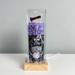 Puede incluir: Lámpara decorativa única con una lata morada "NON STOP" con un gráfico de máscara de gas. La lata está encerrada en una caja de acrílico transparente con un perno oxidado y se asienta sobre una base de madera.