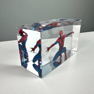 Peut inclure: Un bloc rectangulaire transparent renferme une figurine de Spider-Man dans une pose dynamique. Le super-h&eacute;ros est v&ecirc;tu d'un costume rouge et bleu, les bras tendus. Le bloc refl&egrave;te l'image, cr&eacute;ant plusieurs vues du personnage.
