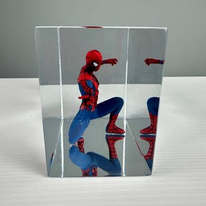 Peut inclure: Une figurine de Spider-Man dans un pr&eacute;sentoir transparent en forme de cube. La figurine, v&ecirc;tue d'un costume rouge et bleu, est en position accroupie. Le pr&eacute;sentoir refl&egrave;te la figurine, cr&eacute;ant un effet miroir. Le fond est gris clair.