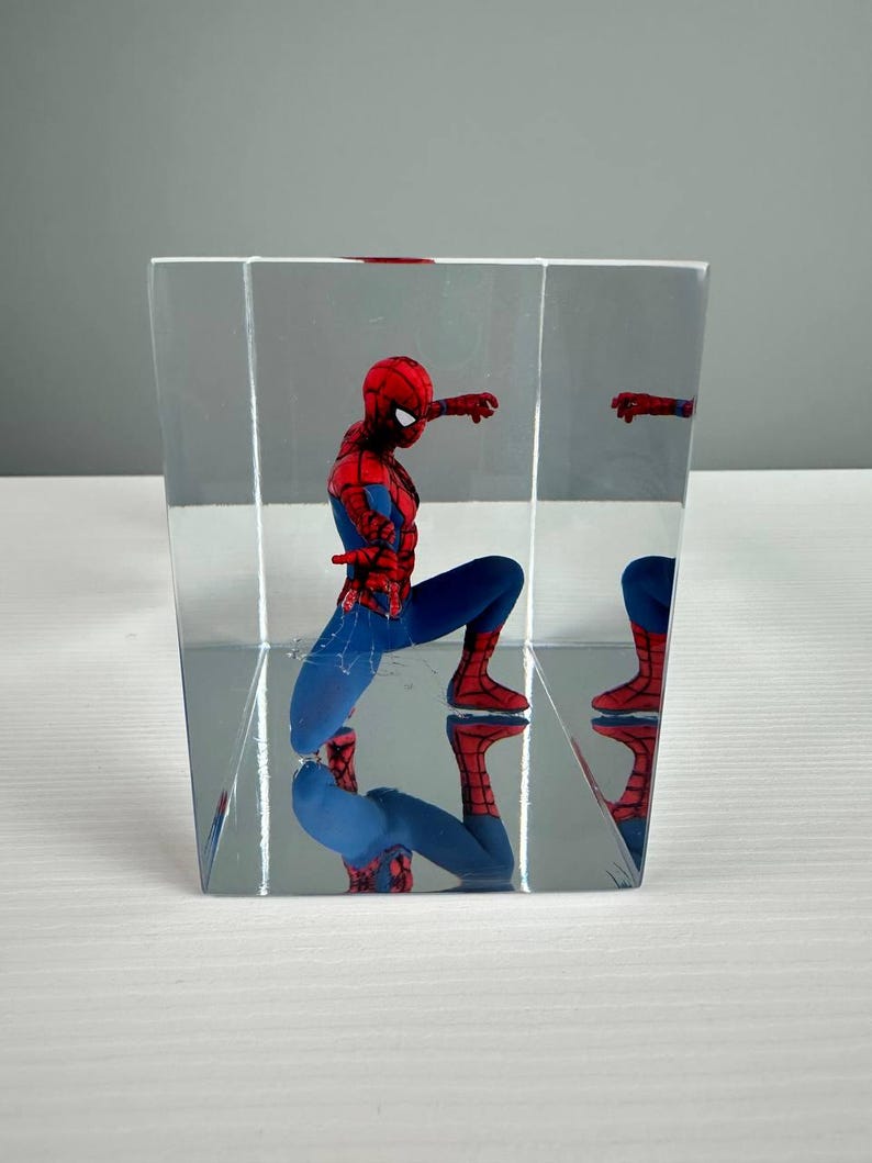 Peut inclure: Une figurine de Spider-Man dans un pr&eacute;sentoir transparent en forme de cube. La figurine, v&ecirc;tue d'un costume rouge et bleu, est en position accroupie. Le pr&eacute;sentoir refl&egrave;te la figurine, cr&eacute;ant un effet miroir. Le fond est gris clair.