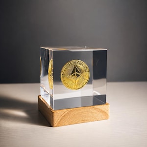 Może przedstawiać: Przezroczysta akrylowa kostka zawierająca złotą monetę Ethereum z widocznym logo i okrągłym tekstem. Kostka jest osadzona na kwadratowej, jasnej drewnianej podstawie. Moneta posiada centralny symbol w kształcie rombu oraz drobne detale.