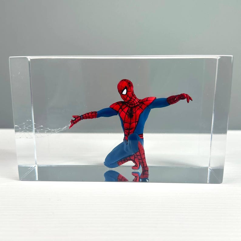 Peut inclure: Figurine de Spider-Man agenouill&eacute;, enferm&eacute;e dans un bloc rectangulaire transparent. Le super-h&eacute;ros porte un costume rouge et bleu, les bras tendus. Le bloc transparent offre une vue claire de la figurine. Le bloc mesure environ 10 cm de haut.