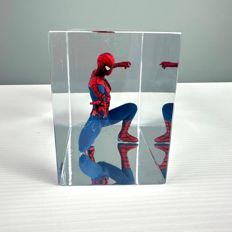 Peut inclure: Une figurine de Spider-Man dans un pr&eacute;sentoir carr&eacute; transparent. La figurine est en position accroupie, v&ecirc;tue d'un costume rouge et bleu. Le bo&icirc;tier refl&egrave;te la figurine, cr&eacute;ant un effet miroir. C'est un objet de collection.