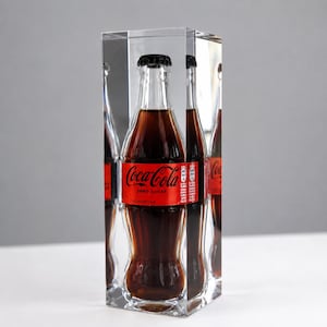 May include: A clear rectangular acrylic block containing a glass Coca-Cola Zero Sugar bottle. The bottle features a red label with the white text Coca-Cola Zero Sugar and a black cap. The transparent block creates multiple reflections of the bottle.