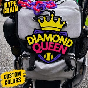 May include: A gray and black baseball equipment backpack with a "Diamond Queen" emblem and a 9-inch chain. The emblem features a yellow crown, a purple and yellow baseball, and the text "Diamond Queen" in yellow and pink. The text "Custom Colors" is also visible.