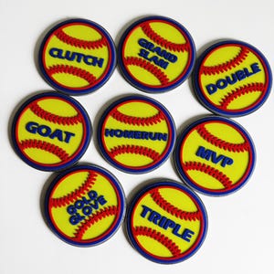 May include: A selection of circular patches with a softball theme. Each patch has a yellow background with red stitching and blue text. The text on the patches includes words such as "CLUTCH," "GRAND SLAM," and "DOUBLE."