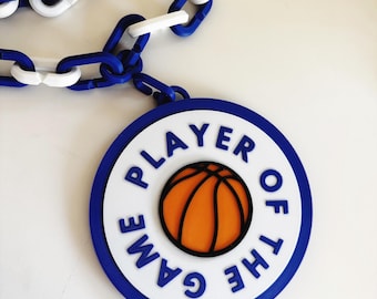 May include: A blue and white basketball-themed Player of the Game award. The round medallion features a basketball graphic and the text "PLAYER OF THE GAME". The award is attached to a blue and white chain.