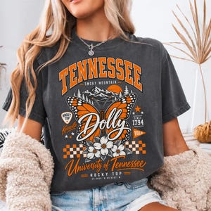 Pode incluir: Uma t-shirt de manga curta cinzento-carvão com uma impressão gráfica com o texto TENNESSEE, Dolly e University of Tennessee. O design inclui uma grande borboleta monarca laranja, flores brancas, silhuetas de montanhas e um padrão xadrez.