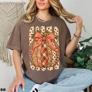 Op de afbeelding: Een bruin t-shirt met korte mouwen en een grote afbeelding van een pompoen versierd met diverse patronen, waaronder luipaardprint, ruitjes en bloemen, bekroond met een grote oranje strik tegen een achtergrond van verticale stippenstrepen.