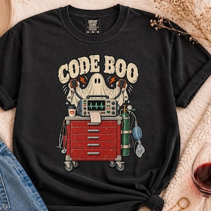 Op de afbeelding: Een zwart t-shirt met korte mouwen en een grafische print van een geest achter een rode medische kar met een hartslagmonitor, zuurstoffles en stethoscoop. De tekst "CODE BOO" staat in een verweerd crèmekleurig lettertype boven de geest.