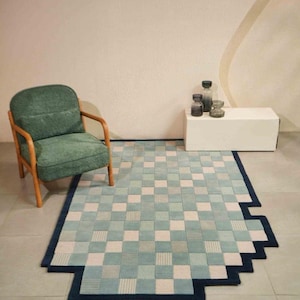 May include: A geometric area rug with a pixelated design in shades of blue, green, and white. The rug has a dark blue border and is placed on a tiled floor. A green armchair with wooden arms sits nearby.