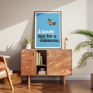 May include: A framed poster with a blue background and the text "A lovely day for a Guinness." The poster features a cartoon toucan. The poster is displayed on a wooden cabinet with a small plant and a wooden hand sculpture.