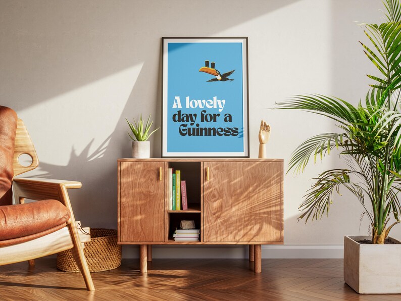 May include: A framed poster with a blue background and the text "A lovely day for a Guinness." The poster features a cartoon toucan. The poster is displayed on a wooden cabinet with a small plant and a wooden hand sculpture.