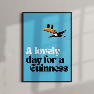 May include: A framed poster with a light blue background features a toucan carrying two pints of Guinness. The text "A lovely day for a Guinness" is displayed in white and black fonts.