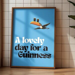 May include: A framed poster with a light blue background features a toucan carrying two pints of Guinness. The text "A lovely day for a Guinness" is displayed in white and black font. The poster is in a brown frame.