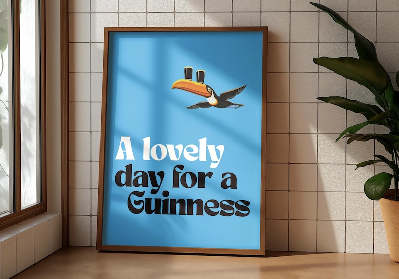 May include: A framed poster with a light blue background features a toucan carrying two pints of Guinness. The text "A lovely day for a Guinness" is displayed in white and black font. The poster is in a brown frame.