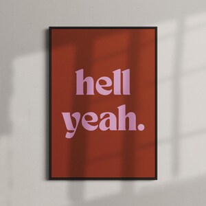 May include: A framed art print with the words "hell yeah." in a pink, bold font. The text is centered on a reddish-orange background. The frame is black, and the print is displayed against a white wall.