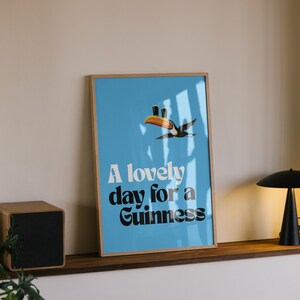 May include: A framed poster with a light blue background and the text "A lovely day for a Guinness." A toucan illustration is above the text. A black lamp and a wooden speaker are on a shelf.