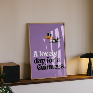 K&ouml;nnte beinhalten: Gerahmtes Poster mit lila Hintergrund und dem Text "A lovely day for a Guinness". Eine Tukan-Illustration befindet sich &uuml;ber dem Text. Eine schwarze Lampe und ein Holz-Lautsprecher sind ebenfalls zu sehen.