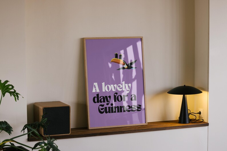 K&ouml;nnte beinhalten: Gerahmtes Poster mit lila Hintergrund und dem Text "A lovely day for a Guinness". Eine Tukan-Illustration befindet sich &uuml;ber dem Text. Eine schwarze Lampe und ein Holz-Lautsprecher sind ebenfalls zu sehen.