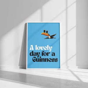May include: Framed poster with a light blue background featuring a toucan carrying two pints of Guinness. The text "A lovely day for a Guinness" is displayed in white and black font.