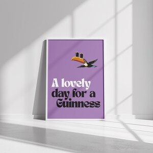K&ouml;nnte beinhalten: Ein gerahmtes Poster mit lila Hintergrund zeigt eine Tukan-Illustration und den Text "A lovely day for a Guinness" in Wei&szlig; und Schwarz. Der Tukan hat einen orangefarbenen Schnabel und ist im Flug.