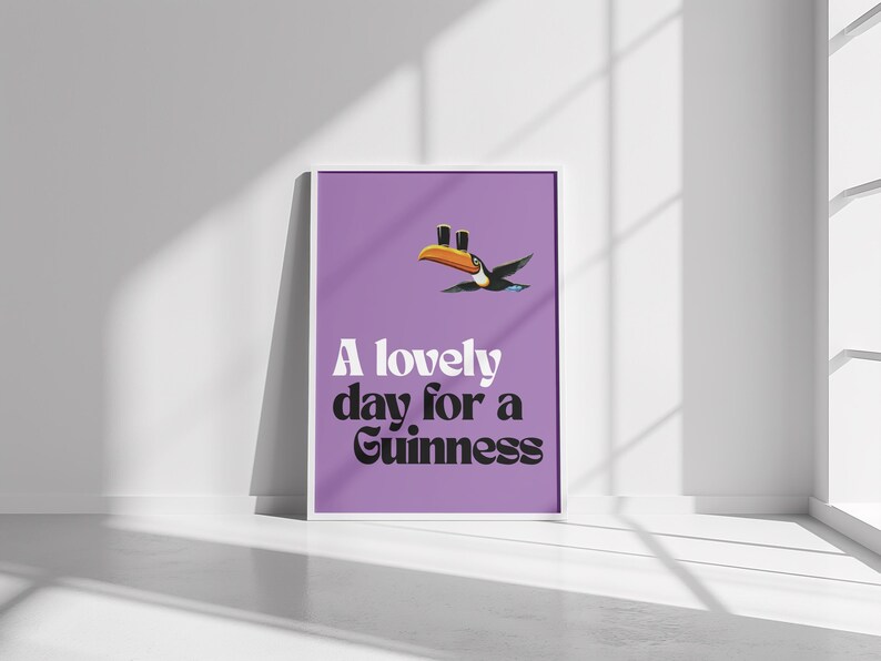 K&ouml;nnte beinhalten: Ein gerahmtes Poster mit lila Hintergrund zeigt eine Tukan-Illustration und den Text "A lovely day for a Guinness" in Wei&szlig; und Schwarz. Der Tukan hat einen orangefarbenen Schnabel und ist im Flug.