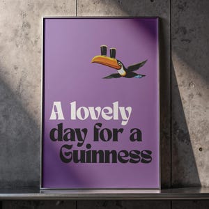 K&ouml;nnte beinhalten: Ein gerahmtes Poster mit lila Hintergrund zeigt einen Cartoon-Tukan mit zwei Bierkr&uuml;gen auf seinem Schnabel. Der Text "A lovely day for a Guinness" ist in wei&szlig;er und schwarzer Schrift dargestellt.
