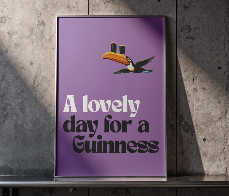 K&ouml;nnte beinhalten: Ein gerahmtes Poster mit lila Hintergrund zeigt einen Cartoon-Tukan mit zwei Bierkr&uuml;gen auf seinem Schnabel. Der Text "A lovely day for a Guinness" ist in wei&szlig;er und schwarzer Schrift dargestellt.