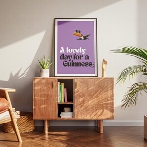 K&ouml;nnte beinhalten: Ein gerahmtes Poster mit lila Hintergrund und dem Text "A lovely day for a Guinness". Eine Tukan-Grafik befindet sich &uuml;ber dem Text. Das Poster wird auf einem Holzschrank mit einer Pflanze und einer Holzhandskulptur ausgestellt.