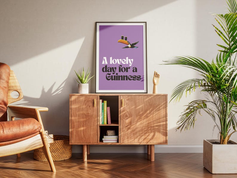 K&ouml;nnte beinhalten: Ein gerahmtes Poster mit lila Hintergrund und dem Text "A lovely day for a Guinness". Eine Tukan-Grafik befindet sich &uuml;ber dem Text. Das Poster wird auf einem Holzschrank mit einer Pflanze und einer Holzhandskulptur ausgestellt.