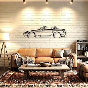 May include: A black metal wall art piece depicting a convertible car silhouette. The artwork is mounted on a white brick wall above a brown leather sofa, a wooden coffee table, and a patterned rug. The room is illuminated by track lighting and a floor lamp.