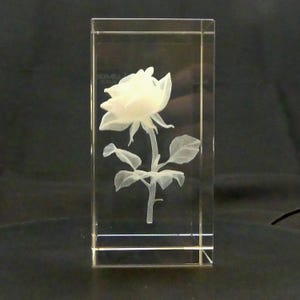 May include: A clear, rectangular crystal block featuring a detailed, three-dimensional carving of a white rose. The rose has delicate petals, leaves, and a stem, all intricately etched within the glass. The block is displayed against a dark background.