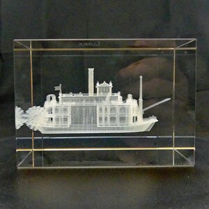 May include: A clear glass block featuring a detailed 3D engraving of a paddle steamer boat. The boat has multiple decks, a smokestack, and a paddle wheel. The block has gold-colored trim and the word "XIMU J" is visible.