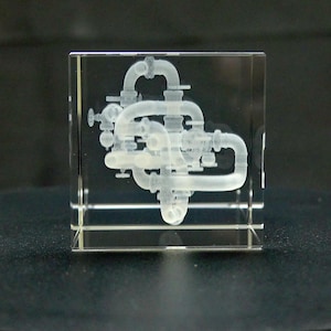 May include: A clear, square glass block with a detailed, three-dimensional rendering of an industrial piping system. The internal structure is a light, translucent color, showcasing the intricate network of pipes, valves, and connectors within the glass cube.