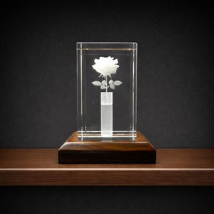 May include: A clear glass block encases a white rose sculpture. The rose is etched within the glass, with detailed petals and leaves. The glass block sits on a dark wooden base, creating a contrast in textures and colors. The overall design is elegant and decorative.