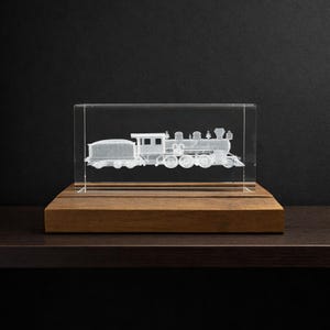May include: A clear glass block with a detailed 3D rendering of a steam train. The train is etched inside the glass, showcasing the locomotive, tender, and passenger cars. The glass block sits on a rectangular wooden base, creating a display piece.