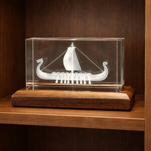 May include: A clear, rectangular glass block encases a detailed, etched image of a Viking longship. The ship features a white sail, oars, and dragon-head prows. It rests on a dark wooden base, displayed on a wooden shelf.