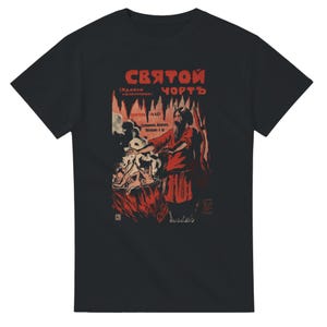 Könnte beinhalten: Ein schwarzes Kurzarm-T-Shirt mit einem Grafikdruck in Rot, Schwarz und Weiß. Das Design zeigt eine gehörnte Figur, die über einem Kessel steht, mit kyrillischem Text "СВЯТОЙ ЧОРТЪ" oben sowie weiterem kleinerem kyrillischem Text.