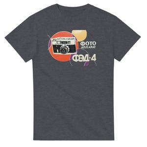 以下が含まれることがあります： ヴィンテージカメラのグラフィックプリントが施されたダークグレーの半袖Tシャツ。デザインには、赤と黄色の円、白でプリントされたキリル文字の「ФОТО АППАРАТ」と「ФЭД-4」、そして細い白い円形のラインアクセントが含まれています。