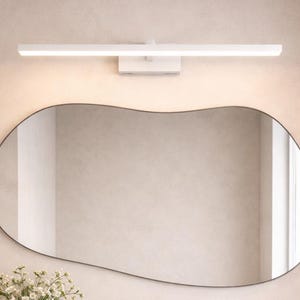 May include: A modern bathroom vanity with a light wood finish, featuring a white countertop, oval mirror, and a white vessel sink. The vanity has two drawers and an open shelf with towels and a basket. A white light fixture is above the mirror.