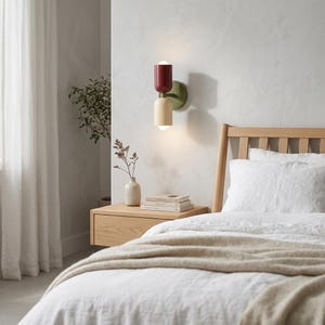 May include: A modern wall sconce with a red top, cream middle, and green base. The sconce is mounted on a light-colored wall above a wooden nightstand. The bedroom setting includes a bed with white linens and a beige throw.