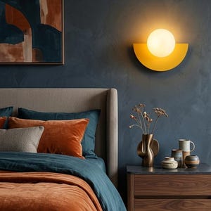 May include: A bedroom scene with a modern aesthetic. A yellow semi-circle wall sconce with a round light is mounted on a dark blue wall. The bed has orange and blue bedding, with a nightstand holding a vase and decorative items.